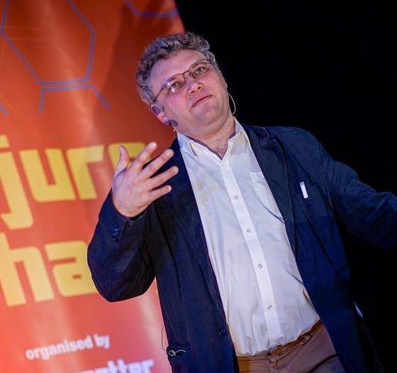 man with curly grey hair and glasses gesturing with hand speaking on stage.