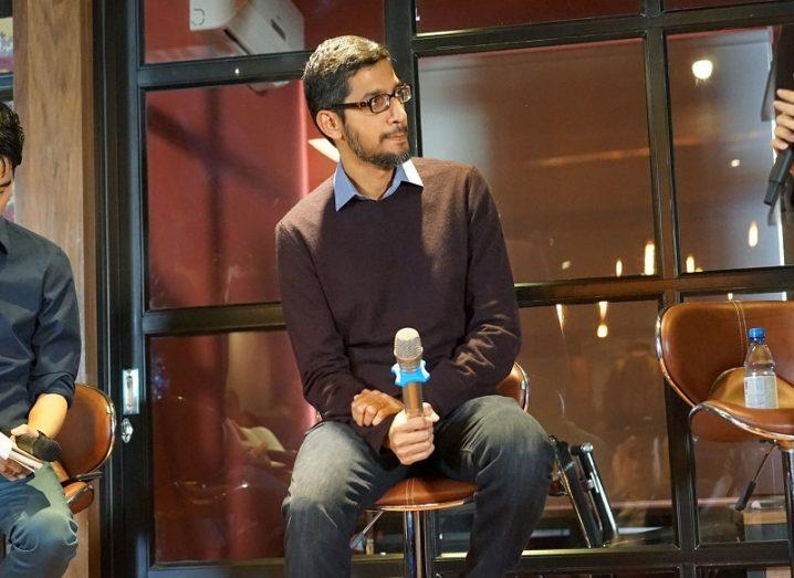 Indian man in brown jumper and blue shirt sitting and holding a microphone during a panel discussion.