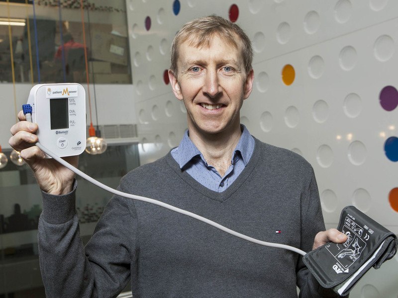 In one hand, a man holds aloft a device connected to a blood pressure monitoring armband in his other hand.