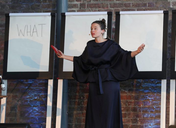 Ada’s Army creator Zoe Philpott stands on stage in front of three flipcharts in front of a brick wall, one chart simple reads: ‘WHAT’