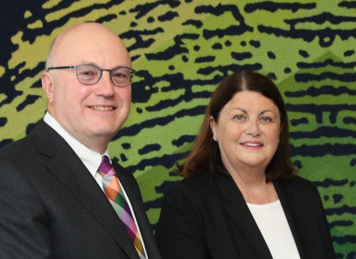 Prof Mark Ferguson and Máire Geoghegan-Quinn stand side by side smiling at the camera in front of a backdrop of a green fingerprint on black ground