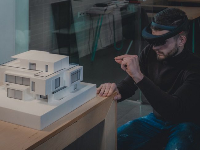 Bearded man wearing an augmented reality headset looking at a 3D model of a house.