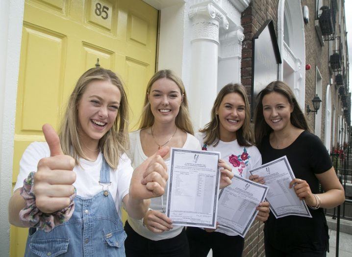 Four smiling students holding their Leaving Cert exam results beside a yellow door.