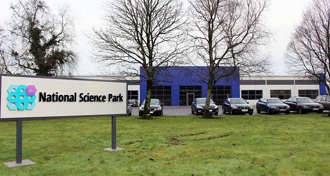 A sign for the National Science Park in a field in the foreground, with the IMR research lab in the background. 