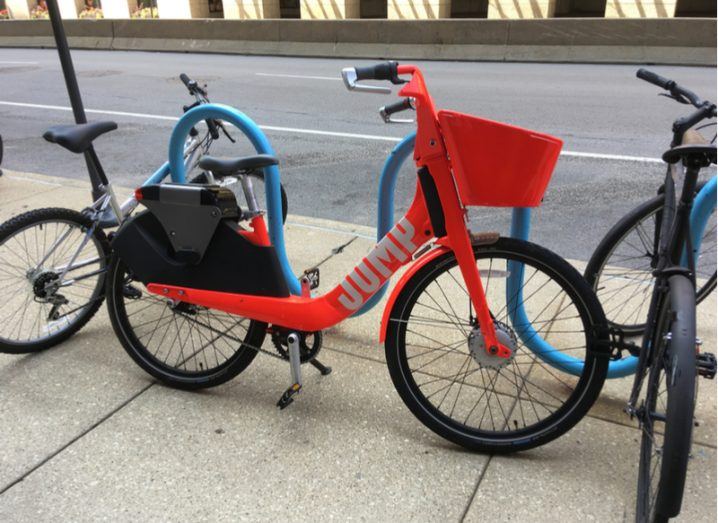 A red Uber Jump ride-sharing bicycle locked to a blue railing on the side of a street.