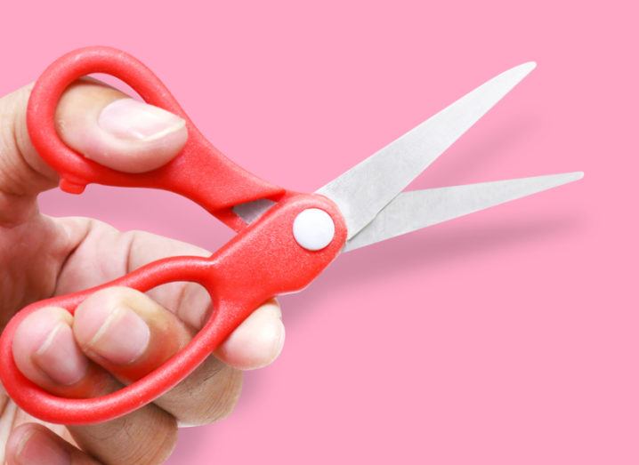 A hand holding a red-handled scissors on a pink background.