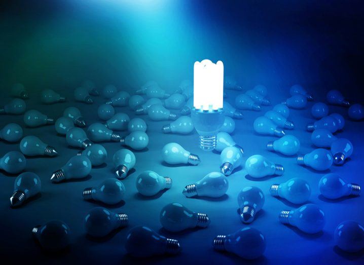 A large glowing lightbulb floats upright amid a blue haze of scattered, unlit small lightbulbs.