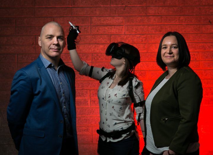 A man and a woman stand beside another woman wearing a VR headset with a red background.