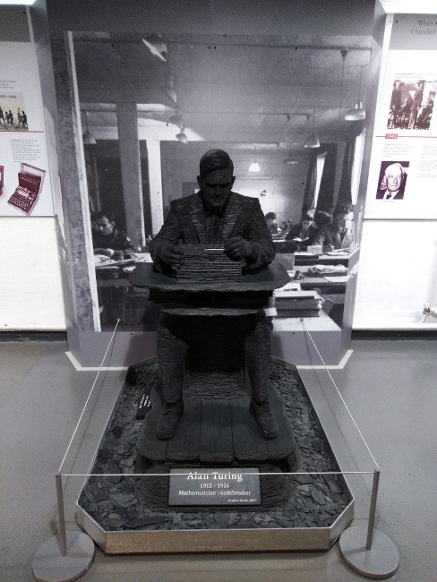 A slate sculpture of a man seated at an Enigma codebreaking machine stands amid a museum exhibition.