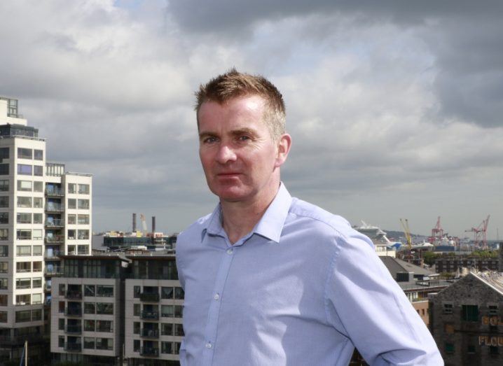 A man in a light blue shirt is photographed on a rooftop with the buildings of Dublin’s docklands in the background.