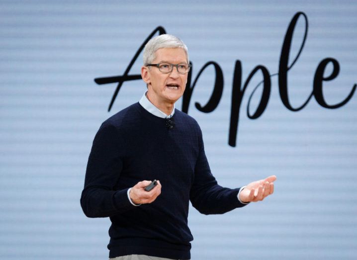 Man in navy jumper presenting on stage in front of blu background with Apple in cursive writing.