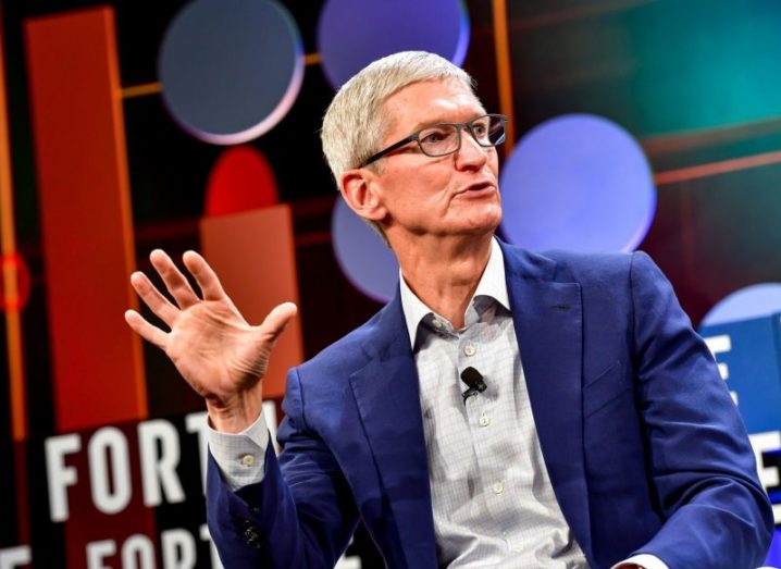 Apple CEO Tim Cook gesticulating at a talk, wearing a navy suit and light grey shirt.