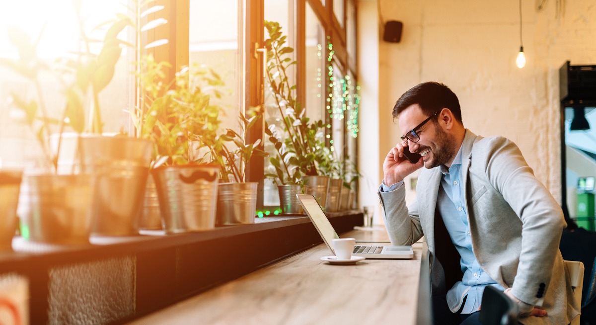 A successful business man talking on the phone while smiling and sitting in front of window.