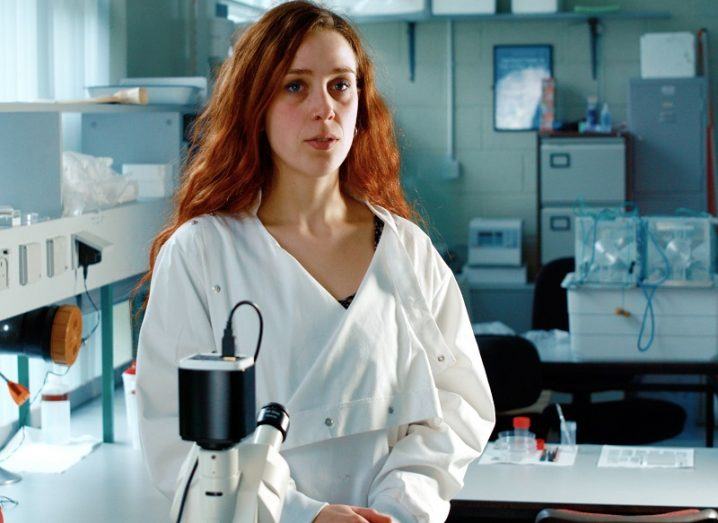 Alina Madita Wieczorek standing in front of a microscope wearing a white lab coat.