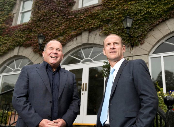 Two men in suits standing before an ivy-covered building.