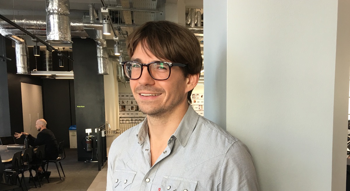 Headshot of a smiling young man with brown hair wearing glasses and an open-neck blue shirt.