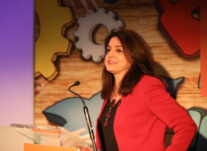 A dark-haired woman in a red blazer stands at a podium in front of a colourful backdrop.