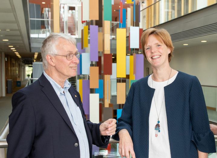 In front of a colourful hanging sculpture, a grey-haired man smiles at a woman with short hair who is looking at the camera.