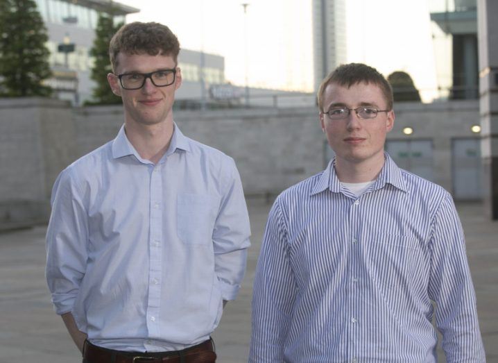 two young men, both wearing glasses and shirts, standing outside.