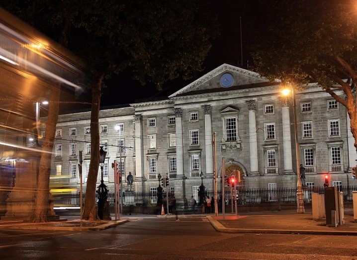 Front of Trinity College Dublin lit up at night with bus appearing into view.