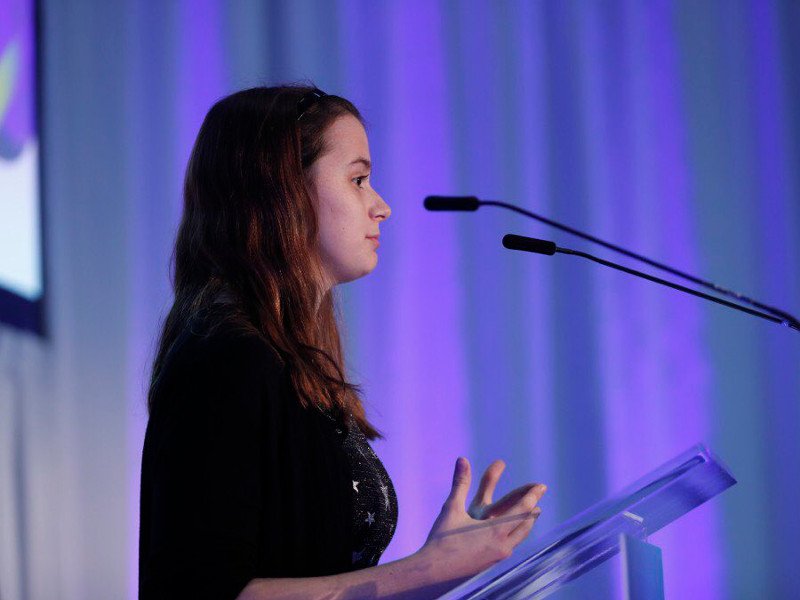 side view of young girl with long brown hair speaking on stage in front of microphone and purple backdrop.