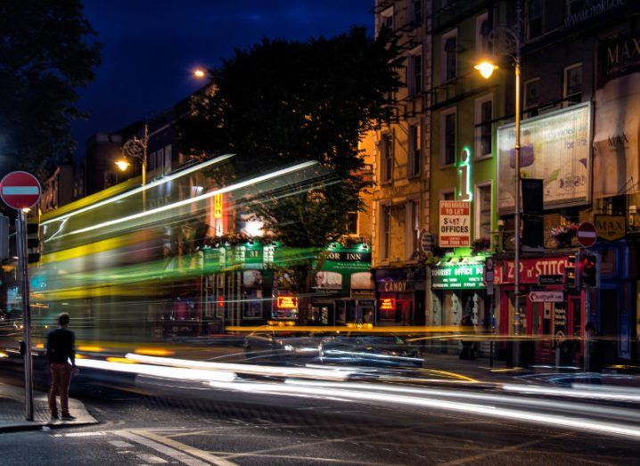 Man stands on Irish street as lights blur past him from traffic.