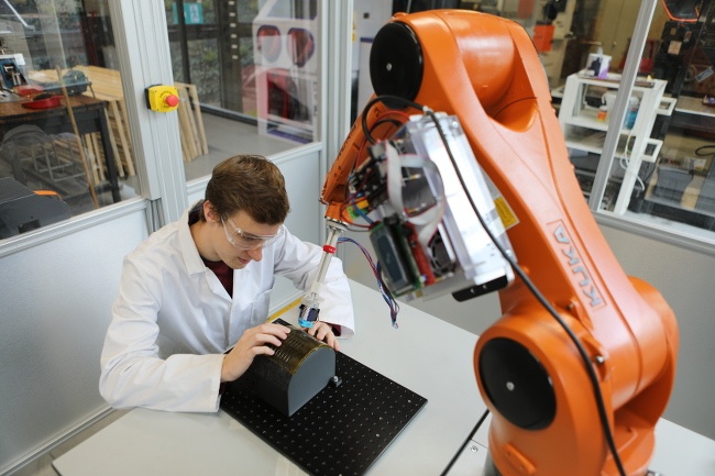 Overhead view of an orange robotic arm printing onto a surface held by a scientist in lab coat and safety glasses.