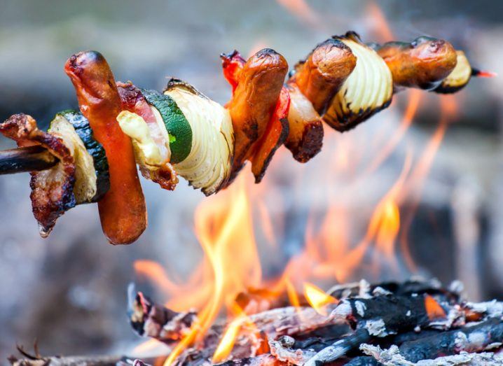 A kebab stick loaded with sausages, courgette slices and onion wedges roasts over an open fire.