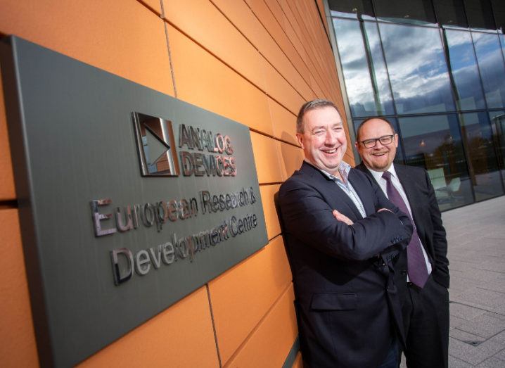 Two middle-aged men in suits, one bald and with glasses, the other with grey slicked back hair, smiling at camera against a backdrop of a plague emblazoned Analog Devices European Research and development centre.