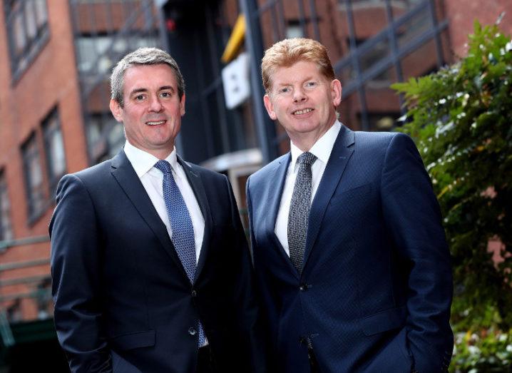 Two men in blue suits stand before a red-bricked building.