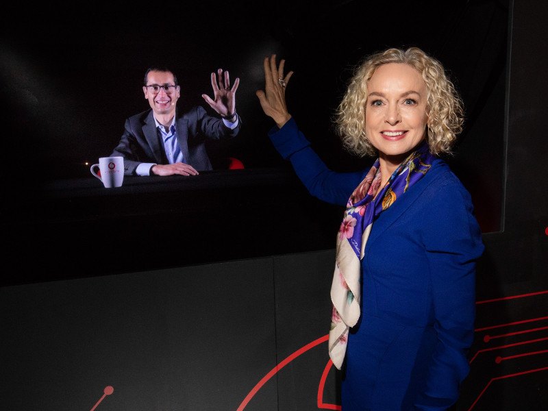 Woman with blonde hair stands before a holographic screen communicating to a man on the screen.