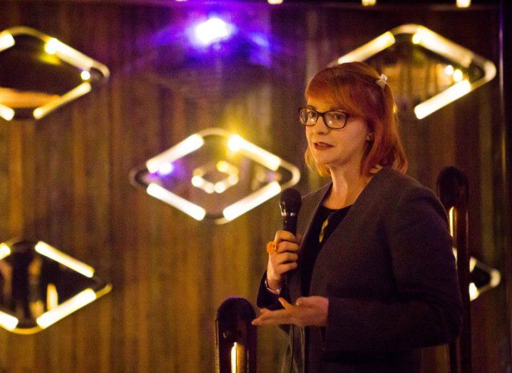 A woman with red hair and glasses gestures while speaking into a microphone in front of a backdrop of diamond-shaped lights.