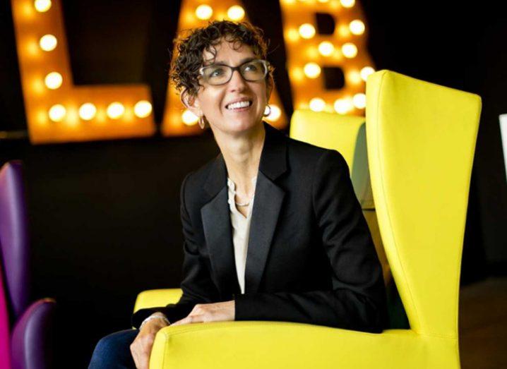 woman in glasses in dark jacket smiles while sitting in a yellow chair.