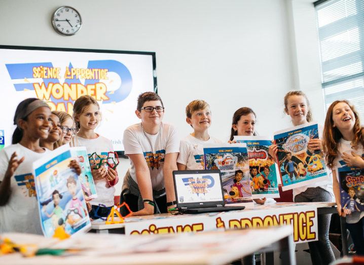 Nine children stand in a row holding up Science Apprentice posters and smiling.