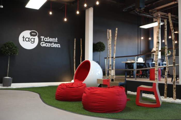 Red beanbags sit on a grass-like floor space. The Talent Garden logo is on the black wall in the background.