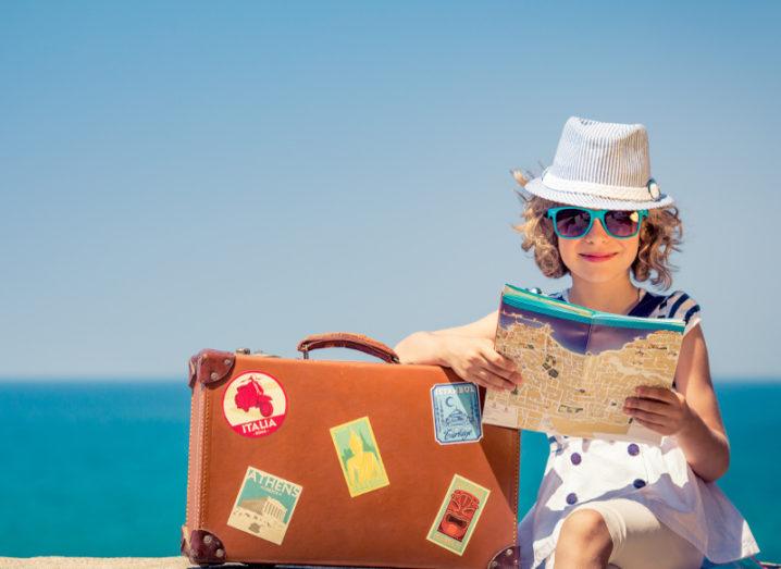 blonde girl wearing sunhat and blue sunglasses reading a map beside a suitcase in front of a blue beach.