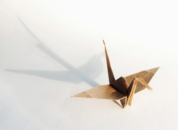 An origami crane made of brown paper casts a long shadow on a white surface.