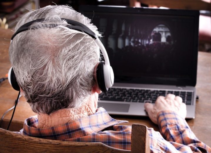 Back of an older man's head wearing headphones as he uses a laptop computer.