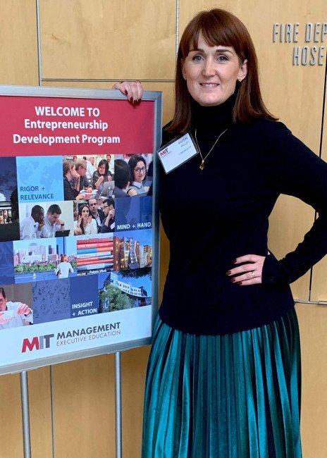 red-haired woman with black polo neck and teal pleated skirt standing beside a welcome sign at MIT.