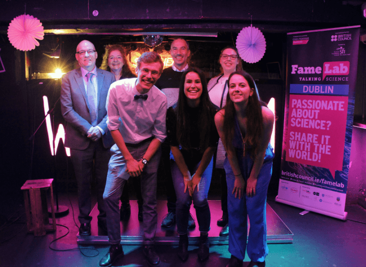 Two rows of people pose for a group photo on the FameLab stage. The three people in front are crouching slightly.