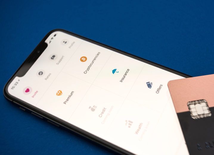 A smartphone screen displays options from the Revolut app menu including ‘Cryptocurrencies’ and ‘Insurance’. A Revolut credit card is placed on top of the phone.