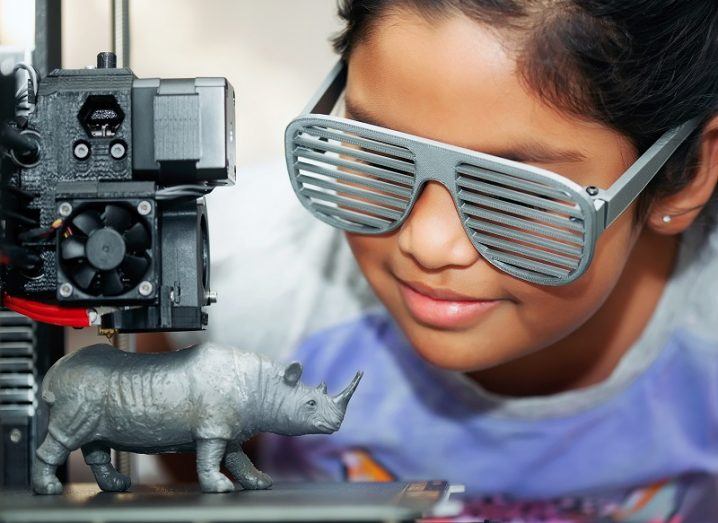 Girl wearing 3D-printed glasses with grill looking at a 3D printer making a small rhino.