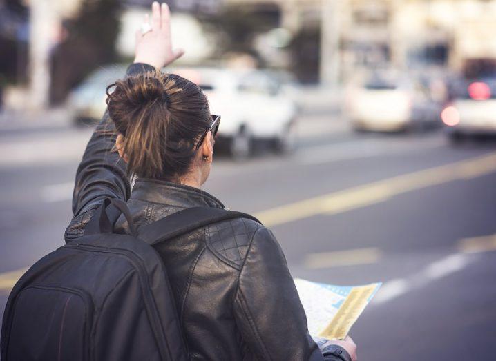 Woman with a black leather jacket and backpack trying to flag a taxi on a busy street.