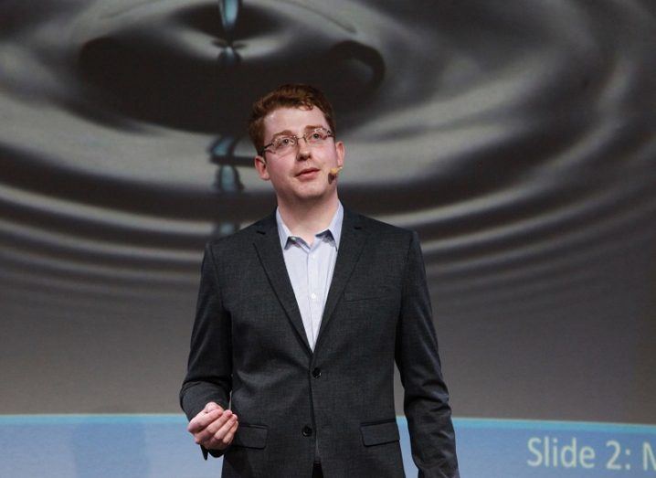 James Blackwell in a black blazer and blue shirt presenting on stage at Threesis, NUI Galway.