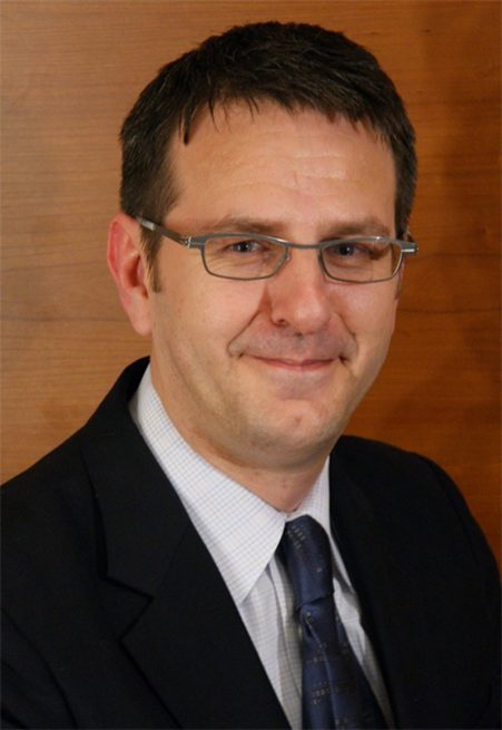 Robin Mersh smiling, wearing glasses and a men's suit jacket with a white shirt and blue tie.