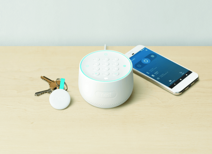 A set of house keys and a smartphone on a table. In the centre, a Nest Guard hub is placed. It is a round, white object with a keypad.