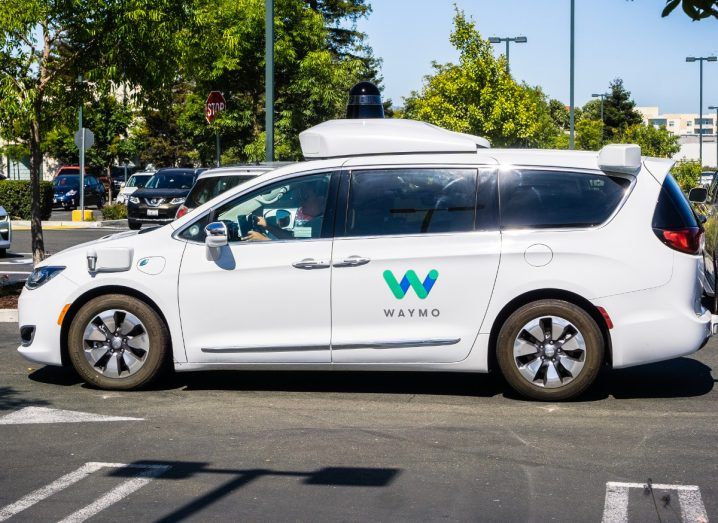 Close up of a white Waymo self-driving car outside on a bright day. The Waymo logo is on the side.