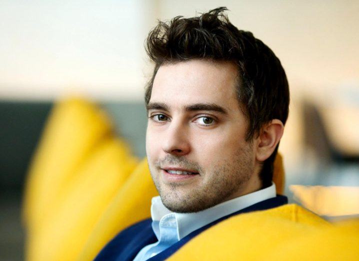 Young man in dark suit with black hair sitting on a yellow-backed chair.