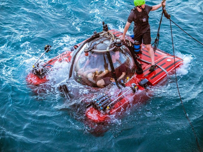 A submersible craft with two-people inside is lowered into the ocean by an operator standing outside the craft and wearing a green helmet.