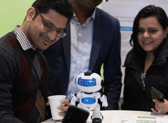 A man holding a coffee cup gets a selfie with a small robot toy while two people wait in the background to take a selfie of their own.